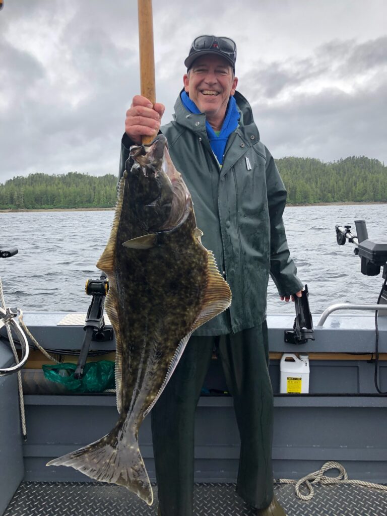 A guy holding a huge halibut with a fish hook and a fishing rig in its mouth.
