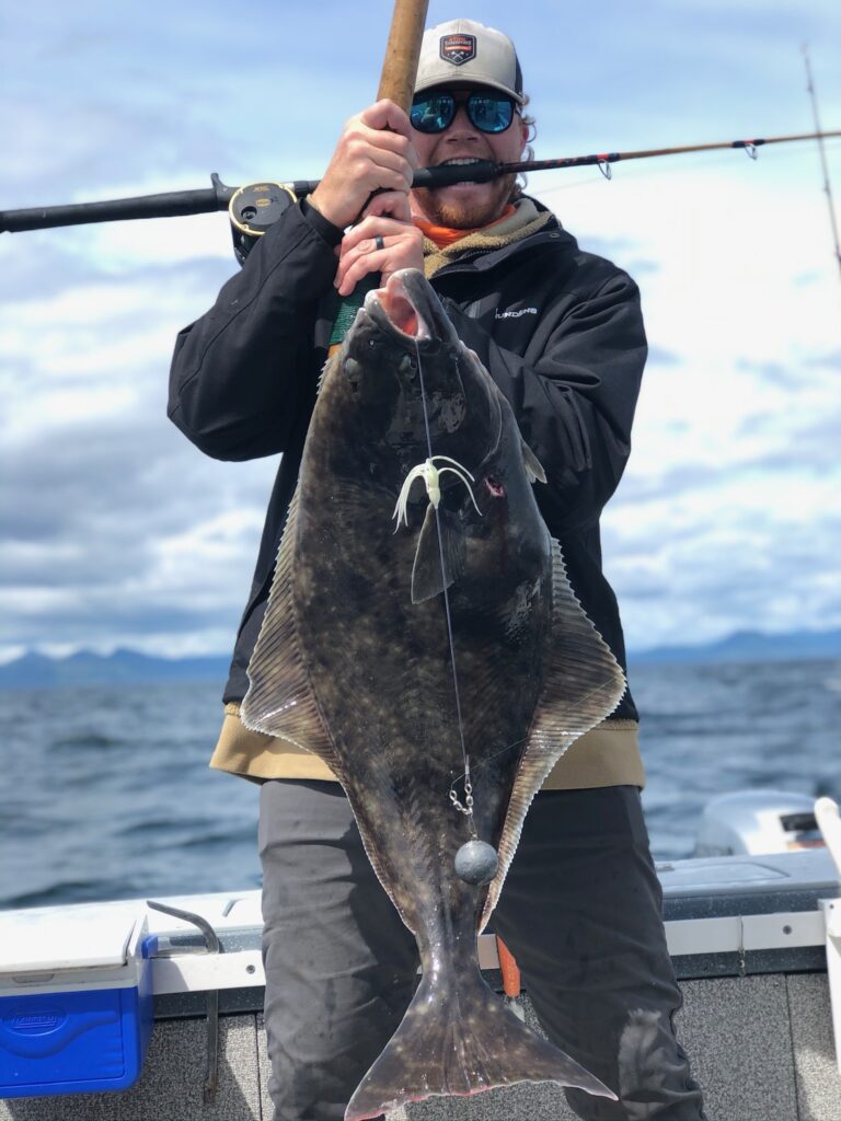 A guy holding a massive pacific halibut with a fishing rig in the halibut mouth and a fishing pole in his mouth.