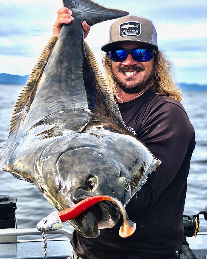 Captain Lukas holding a massive halibut with a fishing rig in its mouth.