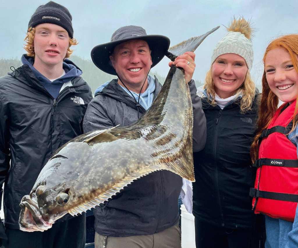 First-time anglers on a fishing charter in Ketchikan holding a massive pacific halibut on a cloudy day.