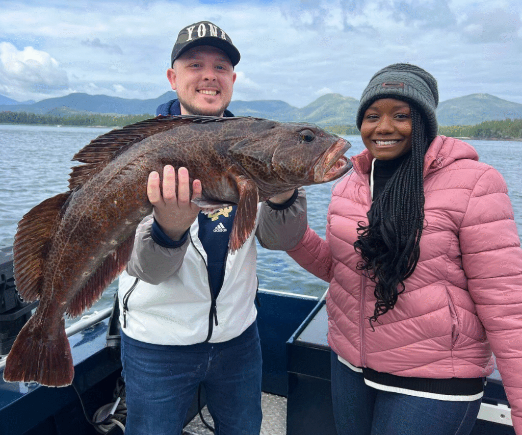 A man and a woman standing on a boat holding a lingcod that was caught during a fishing charter in ketchikan on a cloudy day.