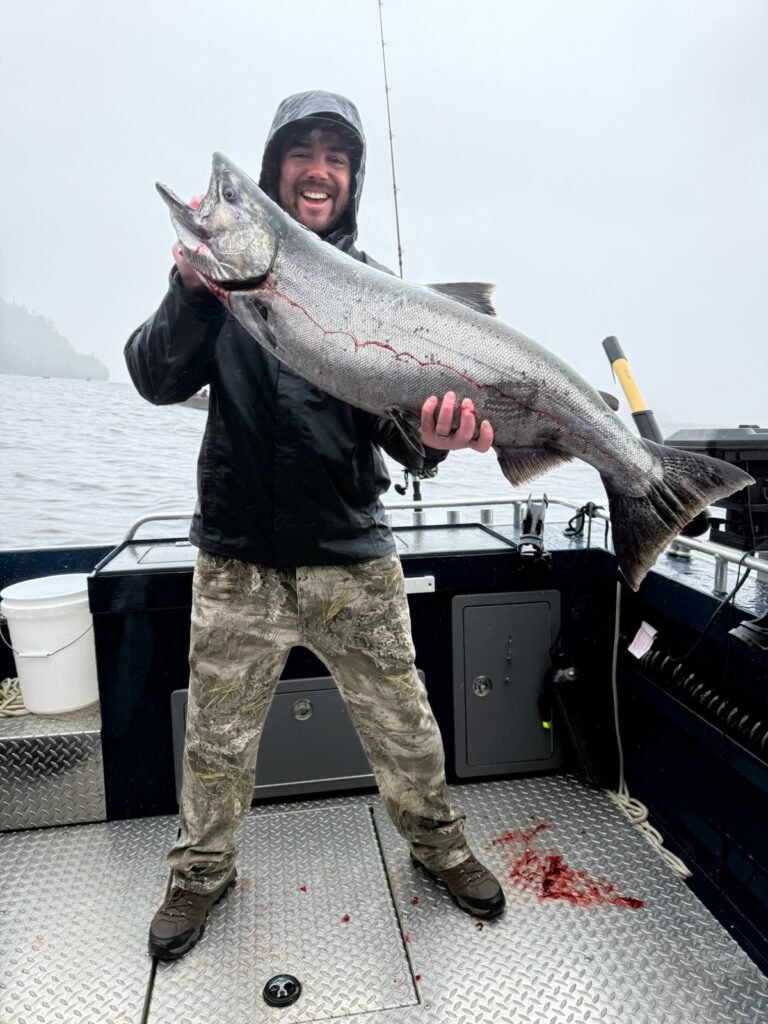 A man holding a massive salmon on a ketchikan fishing charter with rain gear on, hiking boots, and waterproof pants on a rainy day.