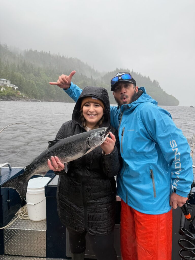 Captain Lukas, of Ketchikan Fishing Company, standing next to a guest with rain gear on holding a salmon during a fishing charter in Ketchikan.