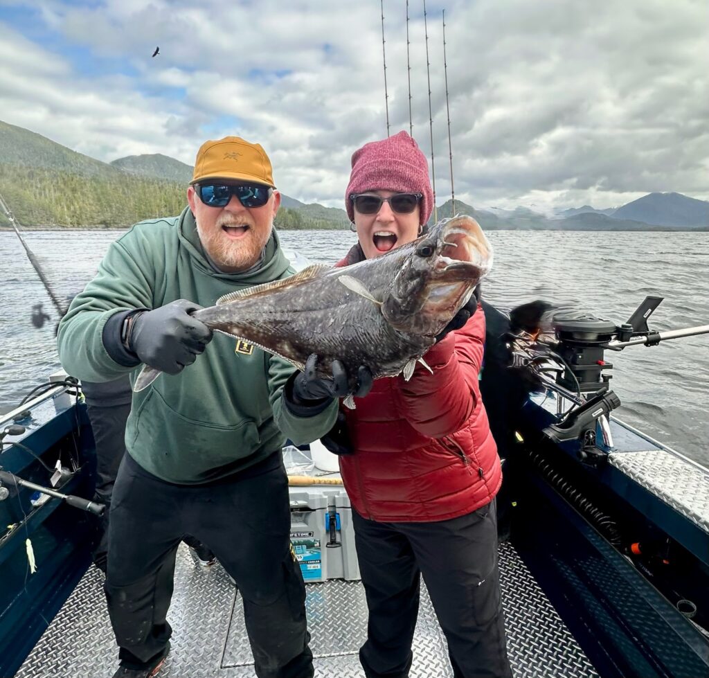 Captain Jake of Ketchikan's Finest Fishing Charters + Ketchikan Fishing Company holding a massive halibut next to a woman on a fishing boat on a cloudy day