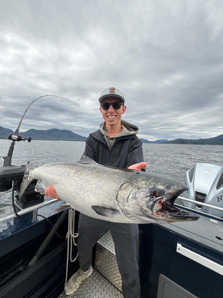A guy standing on a boat on a cloudy day holding a massive king salmon