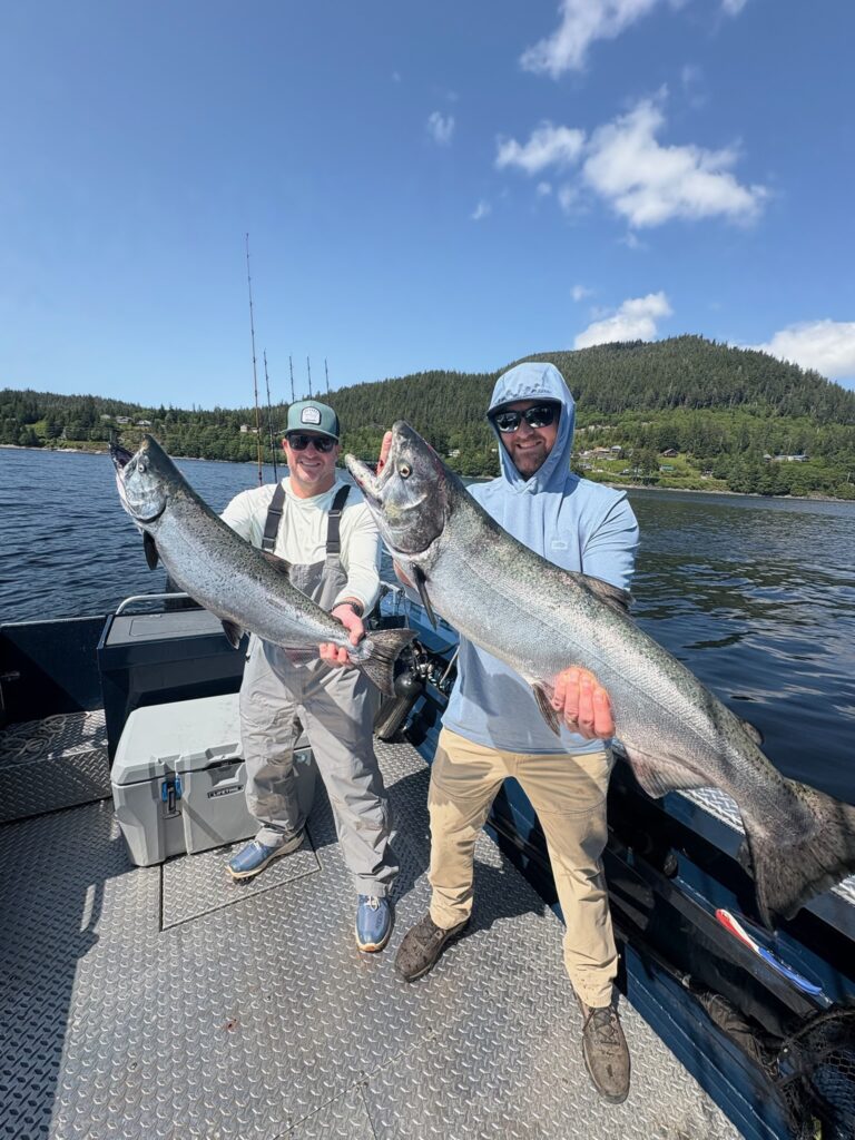 2 guys standing on a ketchikan fishing charter boat on a sunny day holding 2 massive king salmon while wearing waterproof shoes and sunglasses.