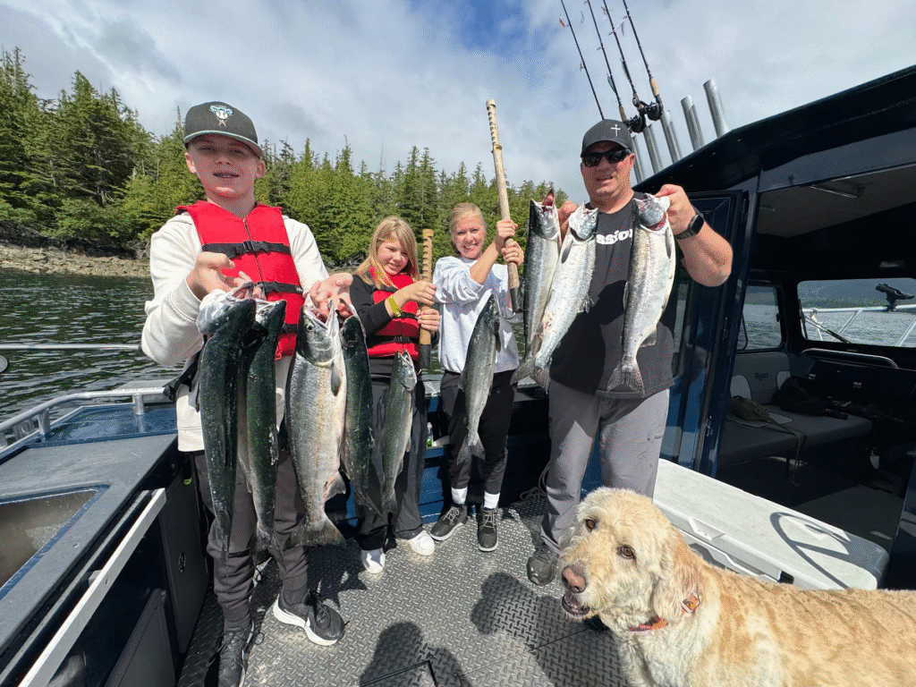 A family smiles while holding a successful salmon haul aboard Ketchikan’s Finest Fishing Charters, with their guide dog Rudy nearby on deck on a sunny day.