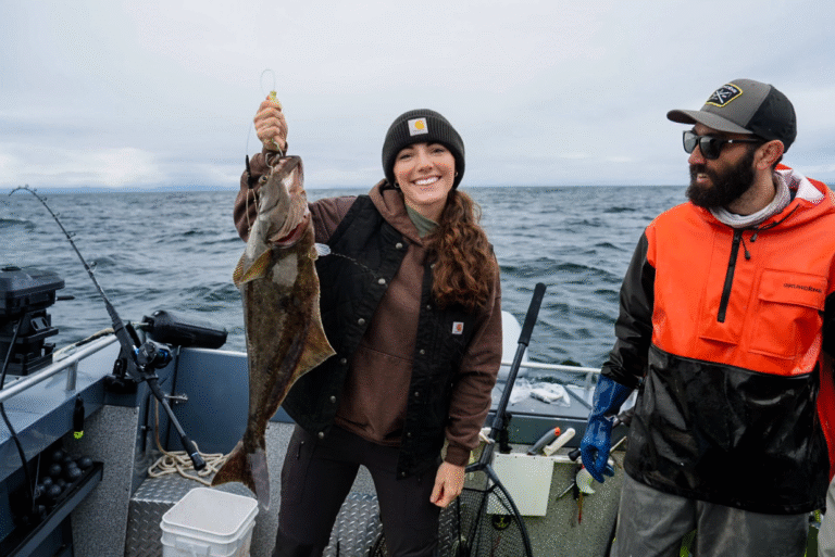 A female guest proudly holds a freshly caught halibut on a cloudy day while smiling on deck with her fishing guide during a Ketchikan charter on her Alaska cruise.