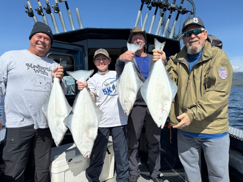 A group of men and women standing on a boat on a sunny day holding massive halibut