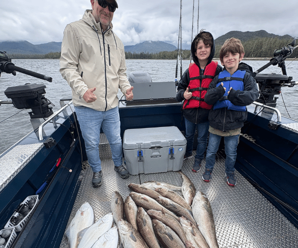 A huge pile of pacific cod and halibut sitting on the deck of a boat with three people standing behind them on a cloduy day