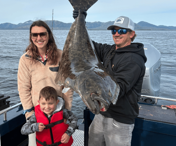 Captain Lukas of Ketchikan Fishing Company holding a huge halibut standing on a boat next to a woman on a cloudy day